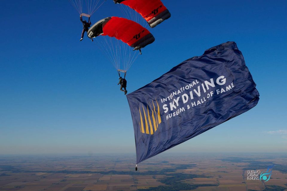 Two skydivers in mid-air hold a large blue flag that reads "International Skydiving Museum & Hall of Fame." They are flying over a vast landscape with clear blue skies.