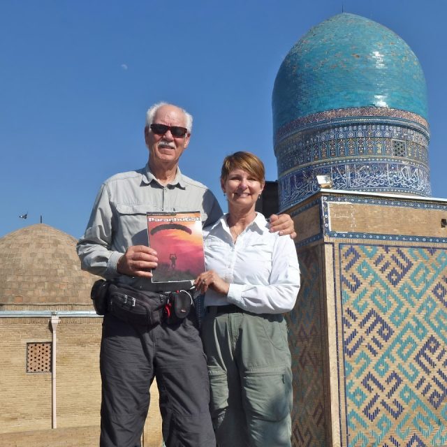 A couple stands in front of a historic building with ornate blue tiles. The man holds a book titled "Paranoia!" Both are wearing casual travel outfits, smiling under a clear blue sky.