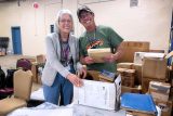 Two people smiling and packing books into boxes on a table in a room full of chairs and more boxes. The woman has gray hair and glasses, and the man is wearing a cap and glasses. They appear to be organizing or setting up for an event.