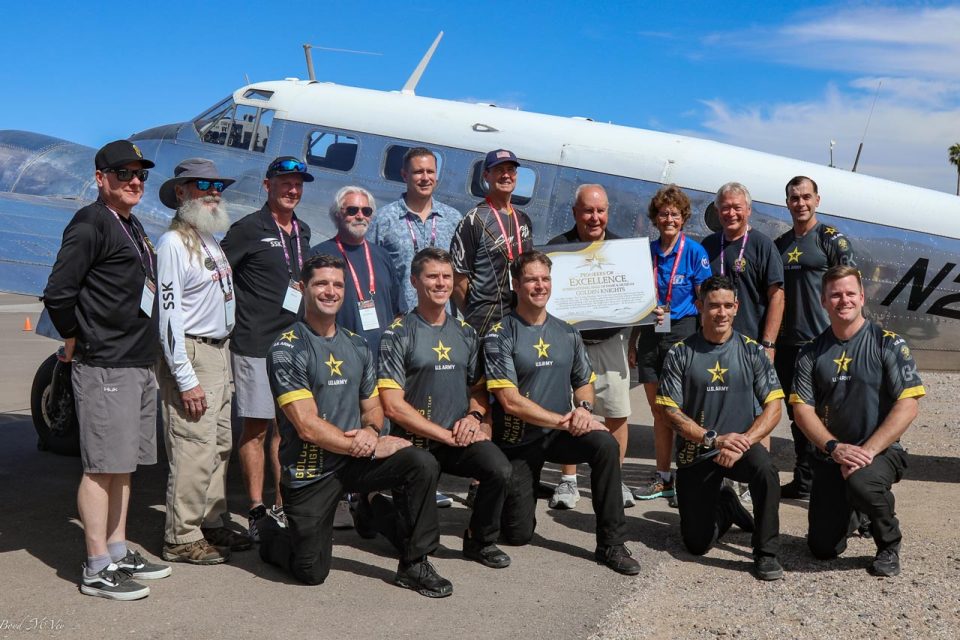 A group of people, including four wearing matching black and gray uniforms, pose in front of a vintage aircraft. One person holds a certificate of excellence. The sky is clear and blue.