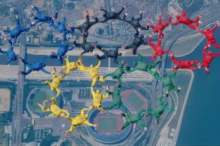 Skydivers in colored suits form the Olympic rings in mid-air over a cityscape with a river and stadium visible below. The formation includes blue, yellow, black, green, and red, representing the Olympic symbol.