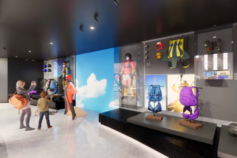 Visitors observe a display of colorful superhero costumes in a museum. Mannequins wear vibrant suits against a backdrop of blue sky and clouds. A woman with a child takes a photo. Bright lighting illuminates the modern exhibit.