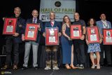 A group of seven people smiling and standing on stage holding framed awards at a Hall of Fame induction ceremony. They are dressed in formal attire, and a banner is displayed behind them.